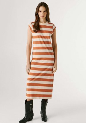 Mujer de pie que lleva un vestido midi sin mangas con rayas horizontales en naranja y beige, y botas negras hasta la mitad de la pantorrilla, contra un fondo blanco.