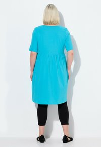 Un vestido turquesa hasta la rodilla con mangas cortas, que presenta una cintura fruncida, combinado con leggings negros y bailarinas negras.