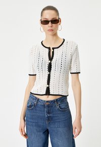 Häkelte weiße Kurzarm-Cardigan mit schwarzem Saum, rundem Ausschnitt, Knöpfen vorne und cropped Passform. Getragen mit hoch taillierten blauen Jeans.