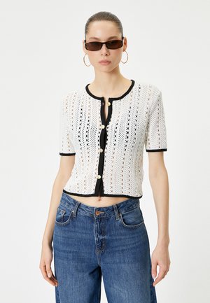 Gehaakte witte cardigan met korte mouwen, zwarte rand, ronde hals, knopen aan de voorkant en een cropped pasvorm. Draagt bij hoge taille blauwe denim jeans.