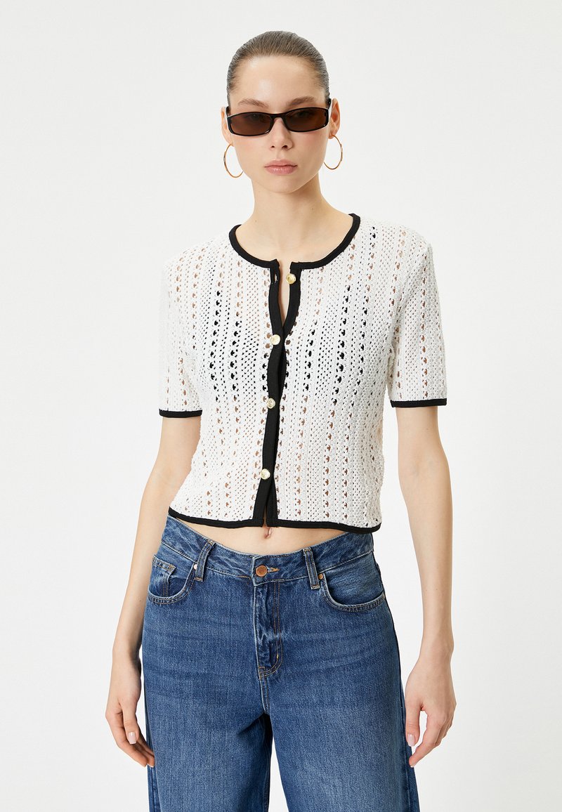 Häkelte weiße Kurzarm-Cardigan mit schwarzem Saum, rundem Ausschnitt, Knöpfen vorne und cropped Passform. Getragen mit hoch taillierten blauen Jeans.