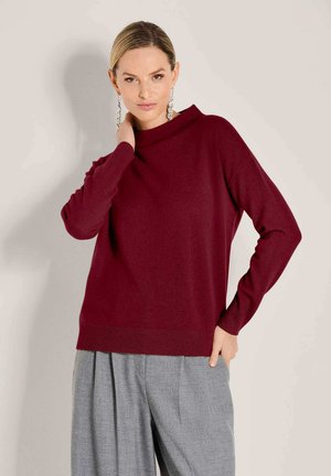 Vrouw in een bordeauxrode trui met lange mouwen en grijze geplooide wijde broek, poseert met één hand op haar nek en de andere in haar zak.