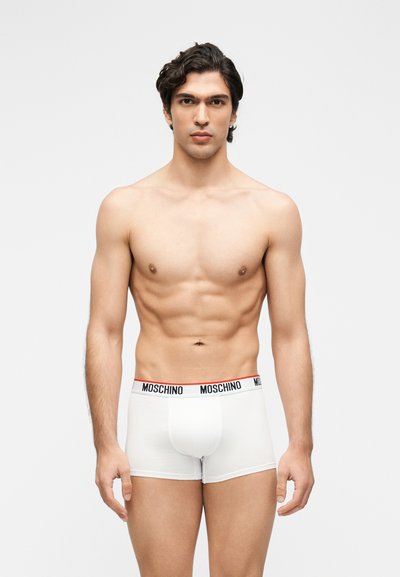 Άνδρας μοντέλο με γυμνασμένο σώμα που φοράει λευκά μποξεράκια Moschino, στέκεται μπροστά σε απλό φόντο ανοιχτού γκρι χρώματος, κοιτάζοντας μπροστά.