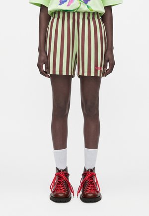 Person iført grønne og brune shorts med lodrette striber, hvide crew sokker og mørkebrune støvler med røde snørrebånd, stående mod en hvid baggrund.