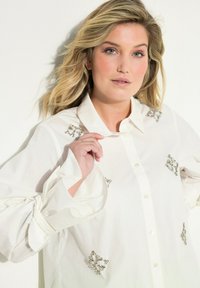 Witte blouse met versierde zilveren accenten. Beschikt over een kraag, knoopsluiting aan de voorkant en gestrikte mouwen. Soepele textuur en een losse pasvorm.