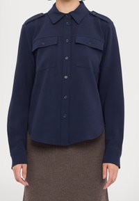 Chemise bleu marine à boutons avec un col pointu, des épaulettes et deux poches poitrine. Fabriquée dans un tissu lisse et doux avec une coupe décontractée.