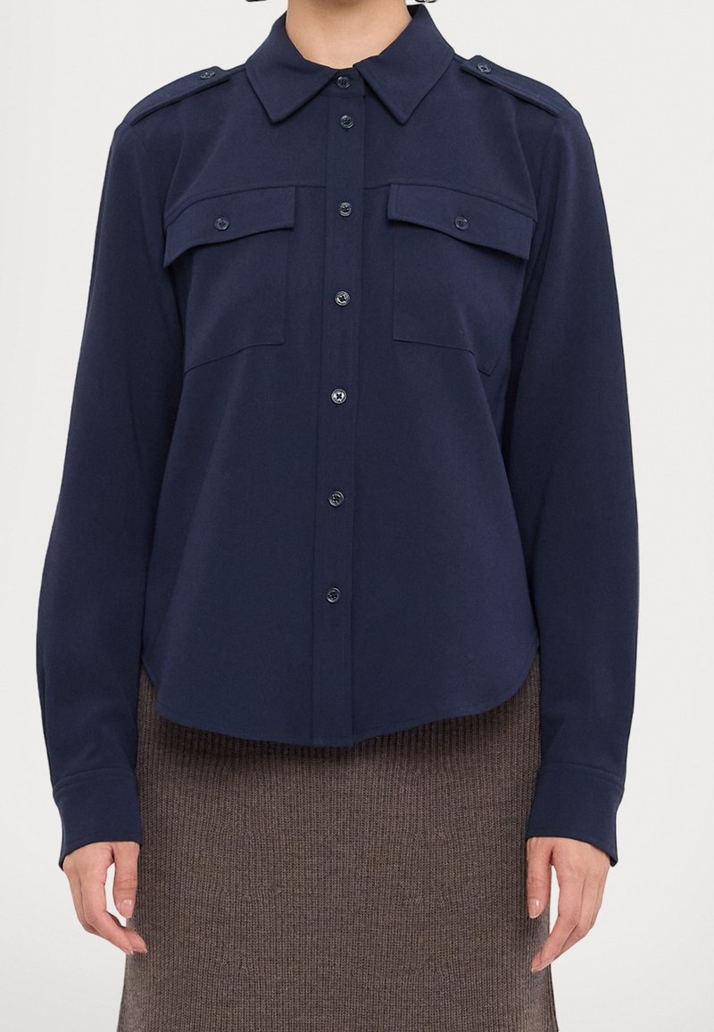 Chemise bleu marine à boutons avec un col pointu, des épaulettes et deux poches poitrine. Fabriquée dans un tissu lisse et doux avec une coupe décontractée.