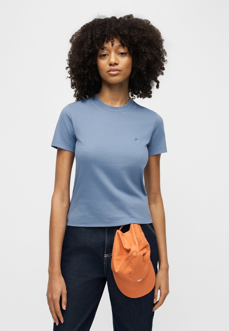 Frau mit lockigem Haar, die ein blaues T-Shirt und dunkle Jeans trägt, hält eine orangefarbene Mütze vor ihrer Taille vor einem weißen Hintergrund.