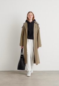 Lungo trench coat kaki sopra un hoodie nero, pantaloni bianchi e sneakers bianche, abbinato a una borsa tote nera. Tessuto morbido, design minimalista.