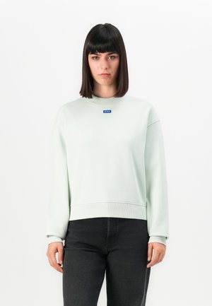 Vrouw met steil donker haar, gekleed in een lichtgroene sweatshirt met het "HUGO" logo en zwarte spijkerbroek, staande tegen een witte achtergrond.