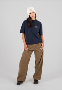 Camiseta oversize de color marino con logotipo dorado, combinada con pantalones de pierna ancha marrones y un gorro de punto crema con un detalle de parche.