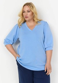 Világoskék V-nyakú blúz puffos ujjakkal, puha anyagból készült. A póló kényelmes fazonú, sima textúrával rendelkezik.