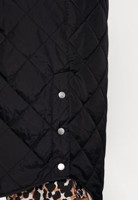 Chaleco acolchado negro con un patrón de diamantes, cierres de botones a presión y una textura suave. El borde inferior es ligeramente alargado.