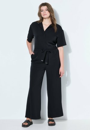 Kobieta stojąca przy białej ścianie, ubrana w czarny kombinezon na krótki rękaw z troczkiem w talii i czarne sandały z odkrytymi palcami.