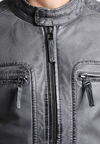 Grå læderjakke med høj krave, lynlås foran, to brystlommer med lynlåse og en diskret tekstureret finish.