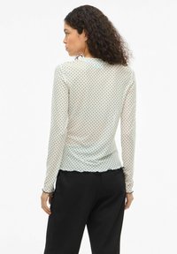 Bluzka w kropki z długim rękawem w jasnokremowym kolorze z czarnymi kropkami, z falowanym dołem i dopasowanym fasonem. Noszona z czarnymi spodniami.