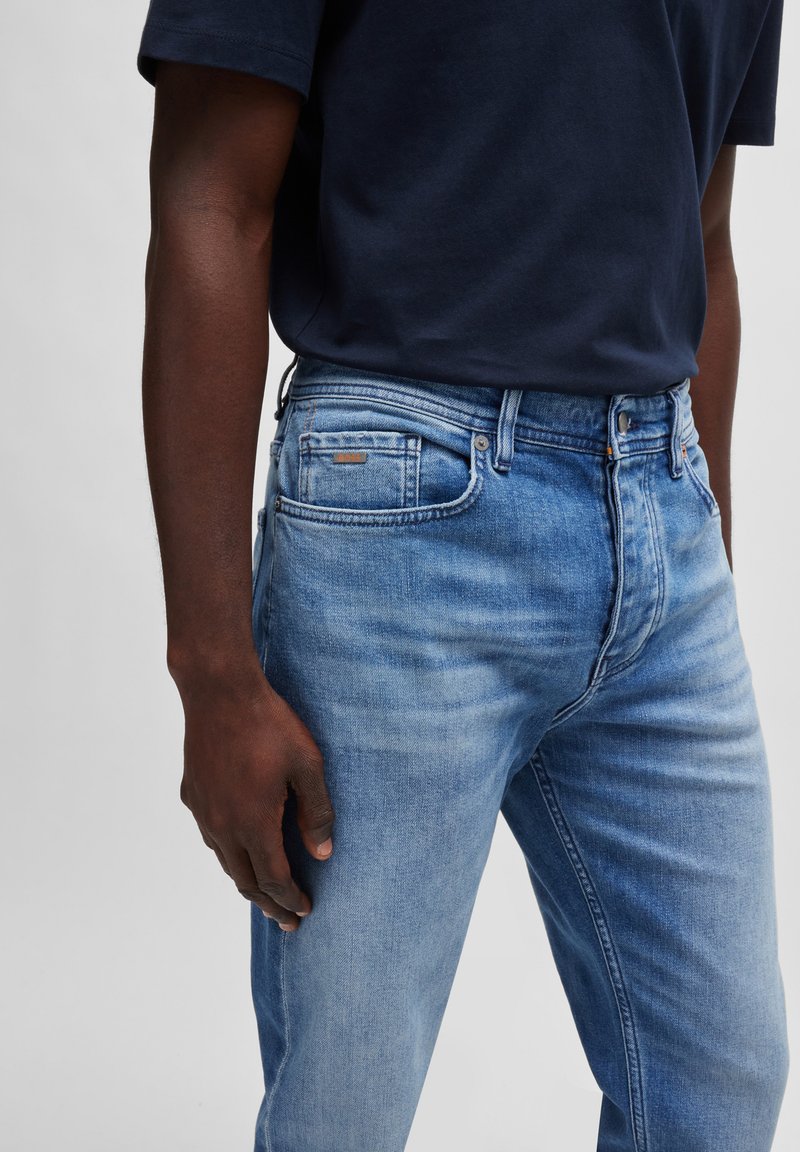 Jean en denim bleu clair avec une texture délavée, cinq poches et une fermeture à bouton. Couture notable et une petite étiquette marron sur la poche arrière.