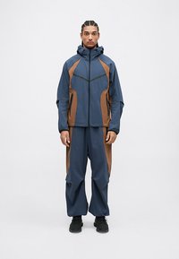 Blauwe buitenjas met bruine accenten, voorzien van een rits aan de voorkant, een capuchon en zijzakken. Losvallende blauwe broek past goed bij de jas.