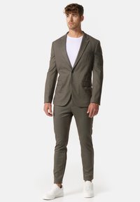 Completo da uomo sartoriale in tessuto a scacchi marrone, caratterizzato da un blazer a un bottone, pantaloni slim-fit e sneaker bianche per un look moderno.