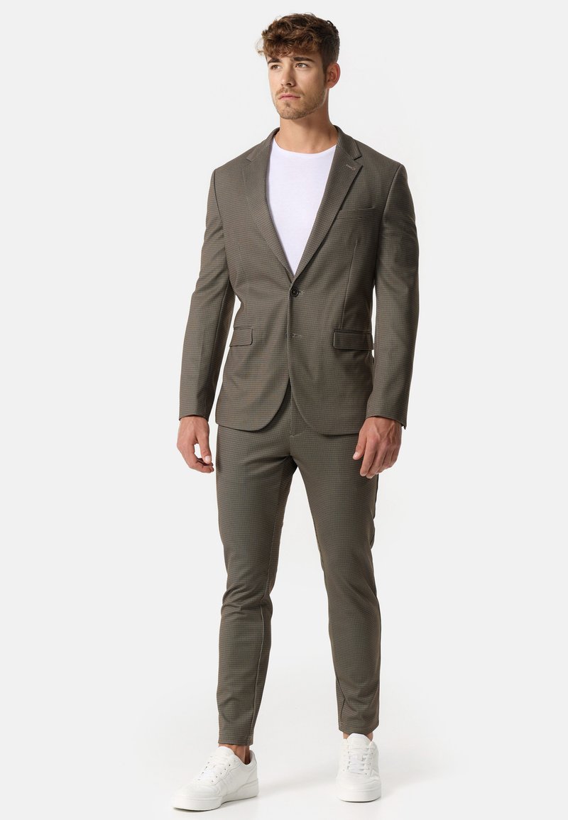 Completo da uomo sartoriale in tessuto a scacchi marrone, caratterizzato da un blazer a un bottone, pantaloni slim-fit e sneaker bianche per un look moderno.