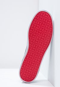 Suola di scarpa rossa con un motivo geometrico, caratterizzata da un bordo bianco lungo il contorno. La suola è esposta in posizione verticale su uno sfondo neutro.