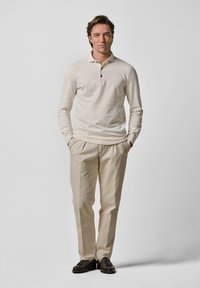 Ljusbeige långärmad polo med tre knappar, kombinerad med krämfärgade byxor och mörkbruna loafers. Enkel, skräddarsydd passform.