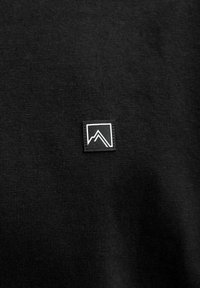 Juodas tekstūruotas audinys su mažo kvadratinio logotipo ženkliuku, baltas kalnų dizainas juodame fone, esantis kairėje pusėje.