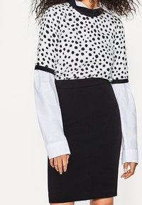 Zwart-witte polkadot top, gelaagd over een witte lange mouw shirt, gecombineerd met een aansluitende zwarte rok. Textuur materialen en contrasterende kleuren.