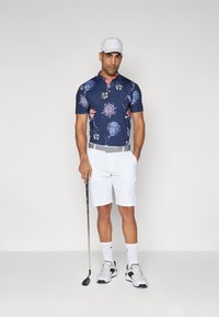 Námorně modré polo tričko s květinovými vzory, červený límec, bílé kraťasy, šedý opasek, drží golfovou hůl, na hlavě má bílou kšiltovku a na nohou tenisky.