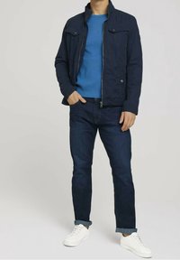 Giacca blu navy con collo alto, maglione blu strutturato, jeans scuri e sneakers bianche. Dotata di tasche frontali e chiusura con cerniera.