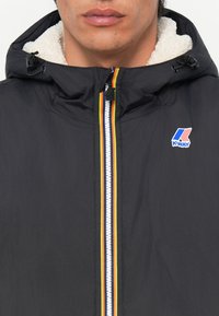 Veste noire avec capuche, dotée d'une doublure en sherpa blanche et d'une fermeture éclair à l'avant avec une bande colorée. Logo K-Way sur la zone de la poitrine.