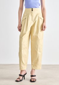 Pantalons beiges taille haute avec des plis, poches avant et deux boutons à la taille. Tissu lisse avec une coupe ample, se resserrant aux chevilles.
