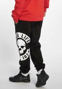 Svart joggebukser med et stort hvitt grafikk som har et hodeskallemotiv og teksten "THUG LIFE 187", laget av myk stoff med en avslappet passform.
