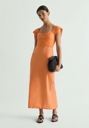 Mujer de pie con vestido midi naranja de manga corta, sosteniendo un bolso de mano negro con cuentas y llevando sandalias negras con tachuelas.