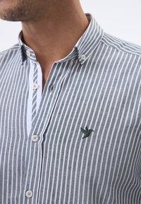 Camisa de botones a rayas en tela gris y blanca. Presenta un pequeño bordado de un pájaro en verde, con un cuello clásico y una fila de botones en el frente.