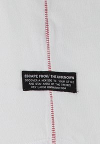 Tela blanca con un visible detalle de costura roja a un lado y una etiqueta negra con texto blanco que dice "Escape From / The Unknown," adjunta en el lateral.