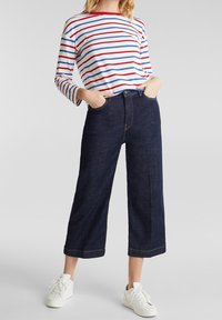 Camicia a maniche lunghe a righe con linee orizzontali rosse, blu e bianche; abbinata a pantaloni ampi in denim scuro e sneakers bianche.