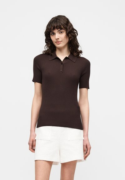 Femme aux cheveux bouclés et foncés portant un polo marron foncé ajusté à manches courtes et un short blanc taille haute avec des poches à l’avant.