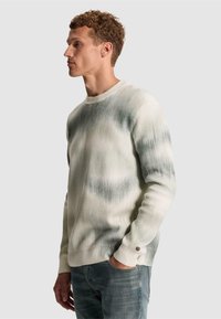 Jeune homme aux cheveux bouclés portant un pull tie-dye blanc et gris et un jean bleu, debout de profil avec une main dans la poche.