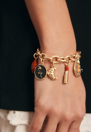 Náramek zlatého řetízku s různými přívěsky: černý kulatý přívěsek s růží, červená kapka, půlměsíc a motiv rtěnky.