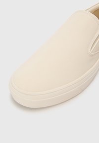 Beige slip-on sko med en glat lædertekstur, rund tå, elastiske sidepaneler og en tyk hvid gummisål. Minimalistisk design.