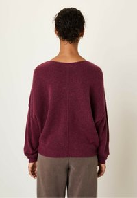 Bordeaux gebreide trui met V-hals, oversized pasvorm, verlaagde schouders en geribbelde manchetten. Gekleed met een paar bruine broeken, gezien vanuit de achterkant.