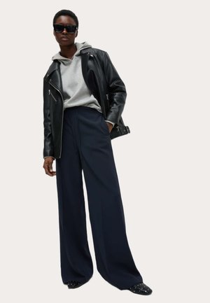 Schwarze Lederbikerjacke über einem grauen Kapuzenpullover, kombiniert mit weiten marineblauen Hosen und schwarzen Slippern mit Nieten. Dunkle Sonnenbrille.