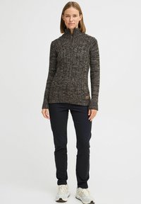 Mørk strikket zip-genser med høy krage, med et teksturert mønster, iført sorte bukser og lyse sneakers.