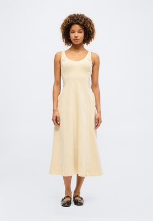 Γυναίκα με σγουρά μαλλιά, ντυμένη με αμάνικο μπεζ μίντι φόρεμα και μαύρα επίπεδα παπούτσια, στέκεται μπροστά σε απλό ανοιχτό φόντο.