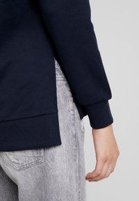 Felpa navy con polsini a costine, fessure laterali e finitura testurizzata, abbinata a jeans grigi chiari strappati.
