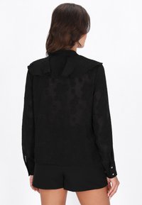 Blusa negra con un patrón floral texturizado, mangas largas y un cuello redondeado con detalle de lazo en la parte posterior. Acabado liso, tela semitransparente.