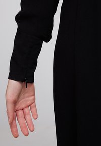 Czarna sukienka z długimi rękawami i dopasowanym mankietem z ukrytym zamkiem. Materiał wydaje się teksturowany, a rękaw ma delikatne marszczenie na ramieniu.