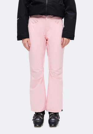 Vue recadrée d’une personne portant un pantalon de ski rose pâle, une veste noire et des chaussures de ski noires sur un fond uni.
