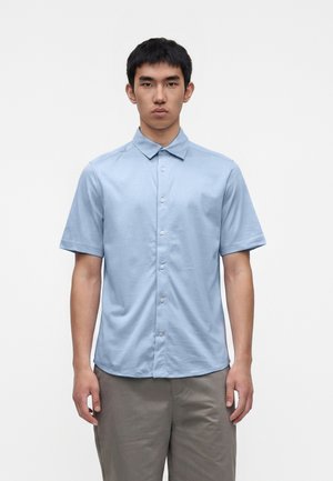 Jeune homme portant une chemise bleu clair à manches courtes et un pantalon gris, debout devant un fond blanc uni, face à l'avant.